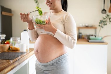 Elinde cam bir kase sebze salatası olan hamile bir kadının fotoğrafını çek ve mutfakta dikilirken afiyetle ye. Doğum öncesi bakım, Hamilelik Diyet yemeği.