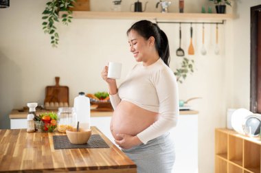 Asyalı hamile bir kadın bardakta su tutuyor ve mutfak tezgahında dururken çıplak karnına dokunuyor. Doğum öncesi bakım, Hamilelik Diyet yemeği.