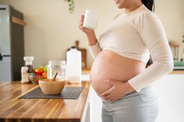 Elinde kupa olan hamile bir kadının mutfak tezgahında durup su içip karnına dokunuşunu yakından çek. Doğum öncesi bakım, Hamilelik Diyet yemeği.