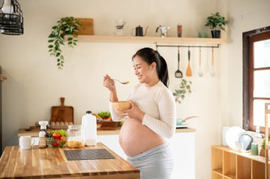 Elinde ahşap bir kâse tutan hamile Asyalı kadın mutfak tezgahında dururken mısır gevreği yemekten zevk alıyor. Doğum öncesi bakım, Hamilelik Diyet yemeği.
