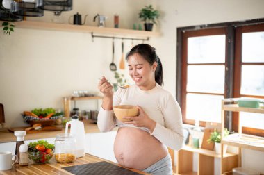 Hamile Asyalı kadın elinde ahşap bir kâseyle gülümserken mutfak tezgahında dikilmiş mısır gevreği yemekten zevk alıyor. Doğum öncesi bakım, Hamilelik Diyet yemeği.