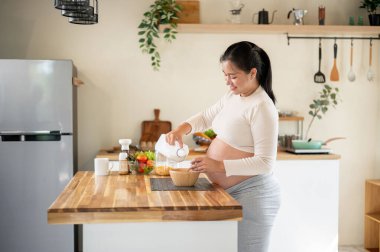 Hamile Asyalı kadın mısır gevreğine süt dökerek mutfak tezgahında kendine yemek hazırlıyor. Doğum öncesi bakım, kendi kendini pişirme, hamilelik diyeti.