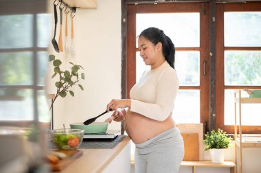 Hamile Asyalı bir kadının mutfakta tavayla yemek pişirişinin yan görüntüsü. Doğum öncesi bakım, kendi kendini pişirme, hamilelik diyeti.