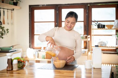 Hamile Asyalı kadın tahıl gevreğini ahşap bir kaseye boşaltıyor ve mutfak tezgahında kendine yemek yapıyor. Doğum öncesi bakım, kendi kendini pişirme, hamilelik diyeti.