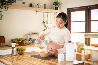 Hamile Asyalı kadın mısır gevreğini ahşap bir kaseye boşaltıyor ve mutfak tezgahında kahvaltı hazırlıyor. Doğum öncesi bakım, kendi kendini pişirme, hamilelik diyeti.