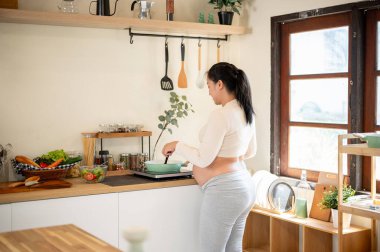 Hamile Asyalı bir kadının, mutfakta tavayla yemek pişirirken görüntüsü. Doğum öncesi bakım, kendi kendini pişirme, hamilelik diyeti.