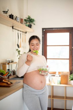 Cam kase tutan hamile Asyalı kadın mutfak tezgahında duran sebze salatasını yemekten zevk alıyor. Doğum öncesi bakım, Hamilelik Diyet yemeği.