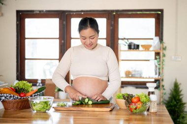 Hamile Asyalı bir kadın mutfakta yemeğini hazırlarken salatalığı bıçakla doğruyor. Doğum öncesi bakım, kendi kendini pişirme, hamilelik diyeti..