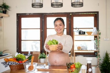 Hamile Asyalı kadın mutfakta taze bir sebzeyi yiyecek malzemesi olarak gösterirken gülümsüyor. Doğum öncesi bakım, kendi kendini pişirme, hamilelik diyeti yemekleri..