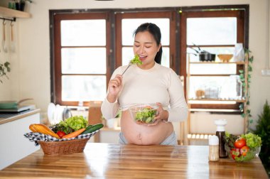 Cam kâse tutan hamile Asyalı kadın mutfak tezgahında yaptığı sebze salatasını yemekten zevk alıyor. Doğum öncesi bakım, Hamilelik Diyet yemeği.