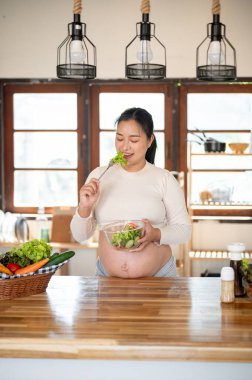 Çatal tutan hamile Asyalı kadın mutfak tezgahında yaptığı sebze salatasını yemekten zevk alıyor. Doğum öncesi bakım, Hamilelik Diyet yemeği.
