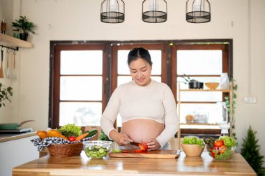 Hamile Asyalı kadın elinde bıçakla domates doğrarken mutfakta salata yapıyor. Doğum öncesi bakım, kendi kendini pişirme, hamilelik diyeti.