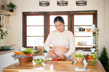 Hamile Asyalı kadın domatesi bıçakla doğruyor. Mutfakta salata yapıyor. Doğum öncesi bakım, kendi kendini pişirme, hamilelik diyeti.