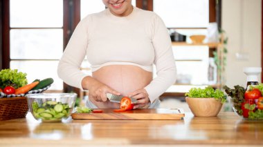 Hamile Asyalı kadının elinde bıçakla domates doğradığı ve mutfakta salata yaptığı yakın plan. Doğum öncesi bakım, kendi kendini pişirme, hamilelik diyeti..