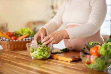 Hamile Asyalı kadının cam kaseye salatalık dilimleri koyup mutfakta sebze salatası yapmasını yakından çek. Doğum öncesi bakım, Hamilelik Diyet yemeği.