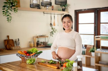 Mutfakta yemek hazırlarken bıçakla salatalık keserken gülümseyen hamile Asyalı kadın. Doğum öncesi bakım, kendi kendini pişirme, hamilelik diyeti..