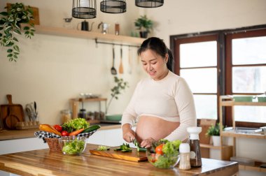Elinde bıçakla salatalık doğrayan hamile bir Asyalı kadın mutfakta yemeğini hazırlıyor. Doğum öncesi bakım, kendi kendini pişirme, hamilelik diyeti..