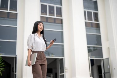 Gömlekli, elinde telefon ve laptop olan Asyalı bir kadın üniversiteden ya da ofis binasından çıkarken önüne bakıyor. Okuldan ayrılan öğrenci, işten çık..