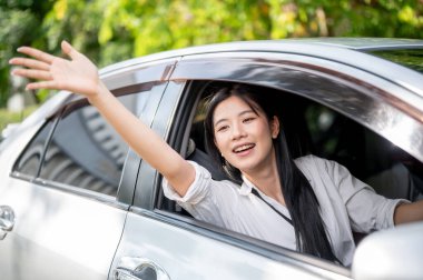 Beyaz tişörtlü Asyalı kadın park yerinde ya da yol kenarında park etmiş bir arabanın sürücü koltuğundan el sallıyor. Arkadaşını almak ya da ailesini beklemek..