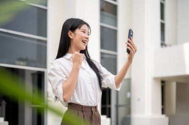 Beyaz tişörtlü Asyalı bir kadın ayakta dururken ya da ofisten çıkarken telefonuna yumruk atıyor. Görüntülü arama ya da kayıt..