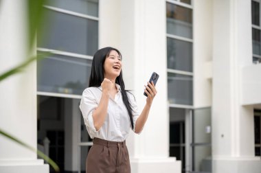 Üniversiteden ya da ofis binasından çıkarken elinde telefon tutan beyaz tişörtlü, gülen Asyalı kadın. Görüntülü arama ya da kayıt..