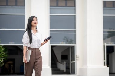 Üniversiteden ya da ofis binasından çıkarken elinde telefon ve laptop olan Asyalı bir kadın. Öğrenci okulu bırakıyor, işçi işten çıkıyor..