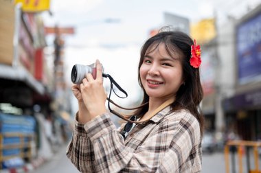 Saçında kırmızı amber çiçeği olan gülümseyen Asyalı turist kadın yerel pazarda yol boyunca kamerayla fotoğraf çekiyor. Yürüyen cadde, Eski kasaba gezisi.