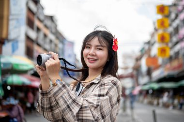 Saçında kırmızı amber çiçeği olan gülümseyen Asyalı turist kadın markette yol kenarında fotoğraf çekiyor. Yürüyen cadde, Eski kasaba gezisi.