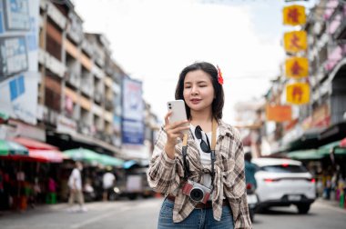 Asyalı turist kadın akıllı telefonları tutuyor ve bakıyor. Yerel bir markette yol kenarında dikilirken yönü kontrol ediyorlar. Yürüyen cadde, Eski kasaba gezisi.