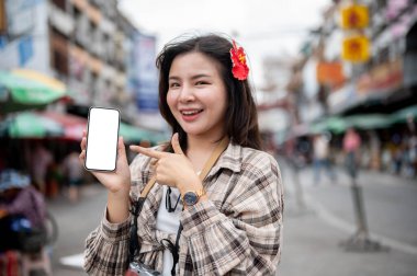 Saçında kırmızı amber çiçeği olan Asyalı turist kadın yerel bir markette beyaz ekran akıllı telefonu gösteriyor ve işaret ediyor. Yürüyen cadde, Eski kasaba gezisi.