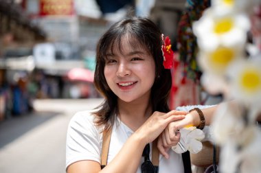 Plastik amber çiçeği tokasıyla gülümseyen Asyalı kadın ve elinde Frangipani bir marketin önünde duruyor. Yürüyen cadde, Eski kasaba gezisi.