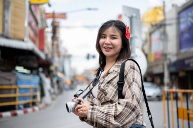 Saçında kırmızı amber çiçeği olan Asyalı turist kadın elinde kamerayla markette yol boyunca fotoğraf çekiyor. Yürüyen cadde, Eski kasaba gezisi.