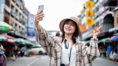 Şapkalı, elinde telefon olan Asyalı bir turist kadın yerel marketin önünde dikilirken bir video görüşmesinde parmağıyla işaret ediyor. Yürüyen cadde, Eski kasaba gezisi.