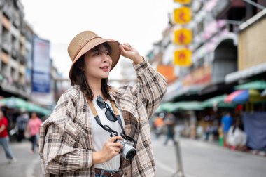 Asyalı turist kadın şapkaya dokunuyor ve elinde kamerayla marketin önünde dikiliyor. Yürüyen cadde, Eski kasaba gezisi.