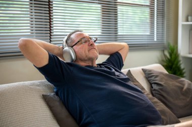 Kafkasyalı, gözlüklü, kulaklı, kafasının arkasına yaslanmış yaşlı beyaz bir adam evdeki kanepede oturuyor. Evde dinlenmek, boş zaman ya da hafta sonu.
