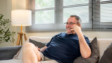 Beyaz, gözlüklü yaşlı beyaz bir adam akıllı telefonla konuşurken gülümsüyor ve evdeki kanepede oturuyor. Evde dinlenmek, boş zaman ya da hafta sonu.