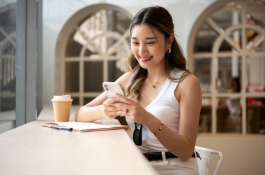 Asyalı bir kadın ahşap masanın üzerinde elinde defter ve kahve fincanı ile telefon tutarken gülümsüyor. Kafede takılıyorum, Kentsel yaşam tarzı.