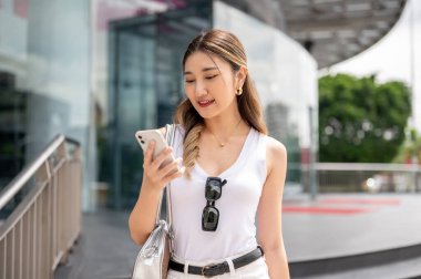 Beyaz kolsuz tişörtlü gülümseyen Asyalı kadın alışveriş merkezinin ön bahçesinde dururken akıllı telefona bakıyor. Dışarıda takılıyorum, para harcıyorum..