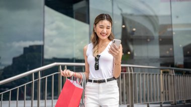 Beyaz atletli Asyalı kadın kırmızı alışveriş çantası tutuyor ve alışveriş merkezinin önünde dikilirken akıllı telefonuna gülümsüyor. Dışarıda takılıyorum, para harcıyorum..