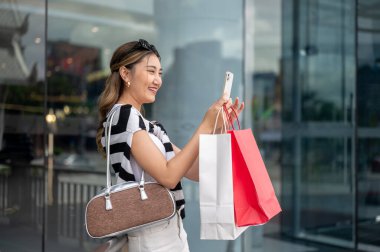 Alışveriş torbaları tutan Asyalı kadın, alışveriş merkezinin önünde korkulukların önünde beklerken telefonla fotoğraf ya da selfie çekiyor. Dışarıda takılıyorum, para harcıyorum..