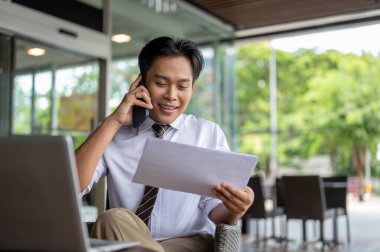 Erkek ofis çalışanı elinde belgelerle kafede ya da kahvecide ahşap masada oturuyormuş gibi telefonla konuşuyor. İş adamı ya da Maaşçı, dışarıda çalışıyor..