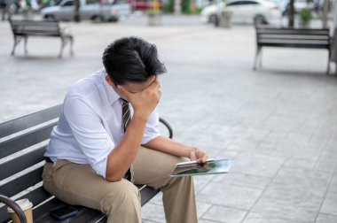 Erkek ofis çalışanı parkta çelik bankta otururken şakağına masaj yapıyor ya da tablet tutuyor. İş adamı ya da Maaşçı, Baş Ağrısı ya da Göz Süzümü.