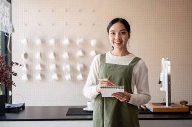 Elinde not defteri olan yeşil önlüklü Asyalı bayan barista gişede müşterinin siparişini almak için bekliyor. Kafe ya da Kafe, Küçük işletme.