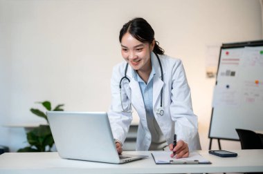 Steteskoplu Asyalı kadın doktor, teşhis odasında dizüstü bilgisayara bakmak için masaya yaslanmış olarak duruyor. Hastane ya da Klinik, Modern ofis.