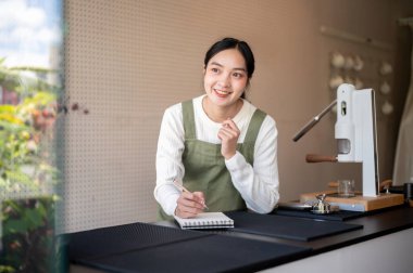 Yeşil önlüklü Asyalı bayan barista tezgahtaki defterde yeni içecek menüleri olduğunu düşünüyor. Kafe ya da Kafe, Küçük işletme.