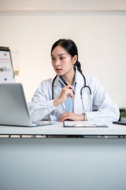 Asyalı kadın doktor çenesinin altında bir kalem tutuyor ve teşhis odasındaki çalışma masasında laptopuna dikkatle bakıyor. Hastane ya da Klinik, Modern ofis.