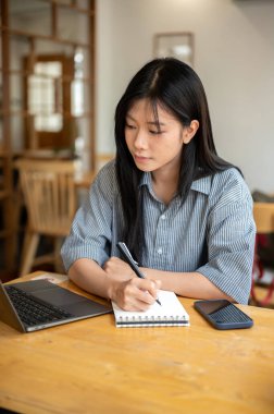 Kafedeki ahşap masada dizüstü bilgisayarına bakarken elinde kalem tutan Asyalı kadın. Çalışıyorum ya da çalışıyorum. Dışarısı Ürpertici.