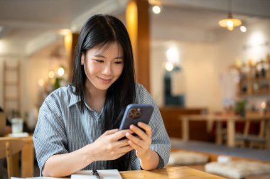 Gülümseyen Asyalı kadın telefonu iki eliyle tutarak video izliyor ya da kafenin ahşap masasında defter üzerinden mesaj yazıyor. Gelir ve Giderler. Para yönetimi.