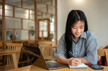 Laptop ve telefonla kafenin ahşap masasına yazı yazarken gülümseyen Asyalı kadın. Çalışıyorum ya da çalışıyorum. Dışarısı Ürpertici.
