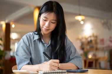 Gülümseyen Asyalı kadın elinde not defteri ve telefonuyla kafenin ahşap masasında kalem tutuyordu. Gelir ve Giderler. Para yönetimi.